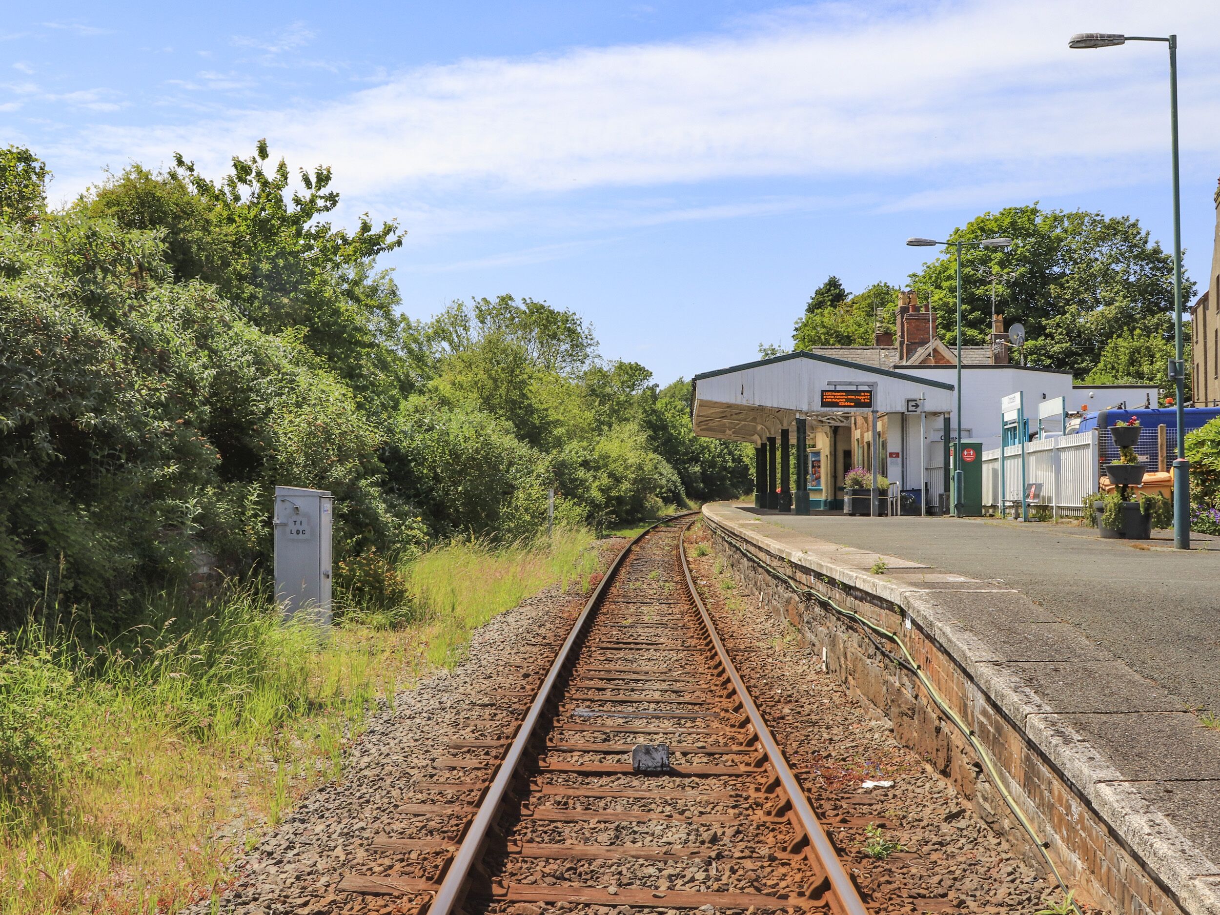 A railway track leading to a station platform with a shelter, surrounded by greenery and a clear sky.