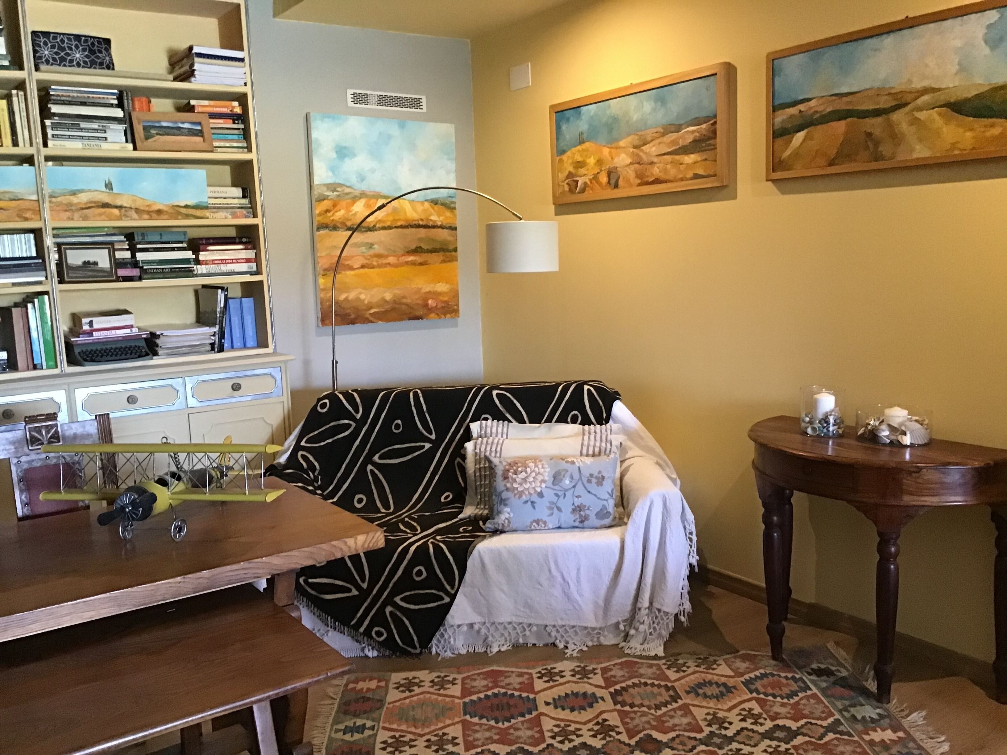 A cozy living room with a patterned sofa, a wooden coffee table, a bookshelf, and two paintings on the wall.