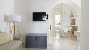 Ein modernes Wohnzimmer mit einem an der Wand befestigten Flachbildfernseher, einem Vintage-Koffer, einer Couch mit Kissen und einem Essbereich mit weißen Stühlen und einem Tisch.