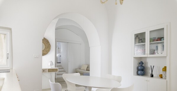 A modern kitchen with a white dining table and chairs, a white cabinet with decorative items, and an arched doorway leading to another room.