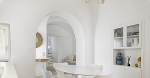 Une cuisine moderne avec une table et des chaises de salle à manger blanches, un meuble blanc agrémenté d’objets décoratifs et une porte en arc donnant sur une autre pièce.