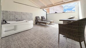 A spa room with a large bathtub, two lounge chairs, a wicker table, patterned floor tiles, and a view of the sky through a window. - Akla Hotel Badajoz (Badajoz)