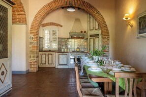 Un espace repas avec une longue table dressée pour le repas, des arches en briques et une cuisine en arrière-plan.