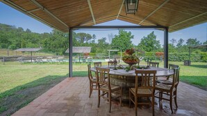 Un espacio cubierto con un patio, una mesa redonda y sillas, un farol, y vistas a una piscina y un jardín.