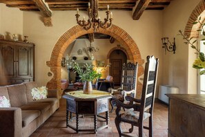 Un salon chaleureux avec une cheminée en briques, une table en bois ornée d’un vase et un canapé agrémenté de coussins à motifs floraux.