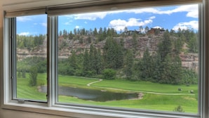 Blick durch ein Fenster auf eine grüne Landschaft mit einem See, Bäumen und einem Gebäude auf einem Hügel.
