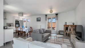 Ein modernes Wohnzimmer mit grauem Sofa, weißen Sesseln, einem Esstisch mit Stühlen, einem Fernseher und einem Kamin.