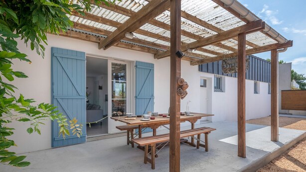 Eine überdachte Terrasse mit einem Holztisch und Bänken, einer weißen Außenwand und blauen Fensterläden.