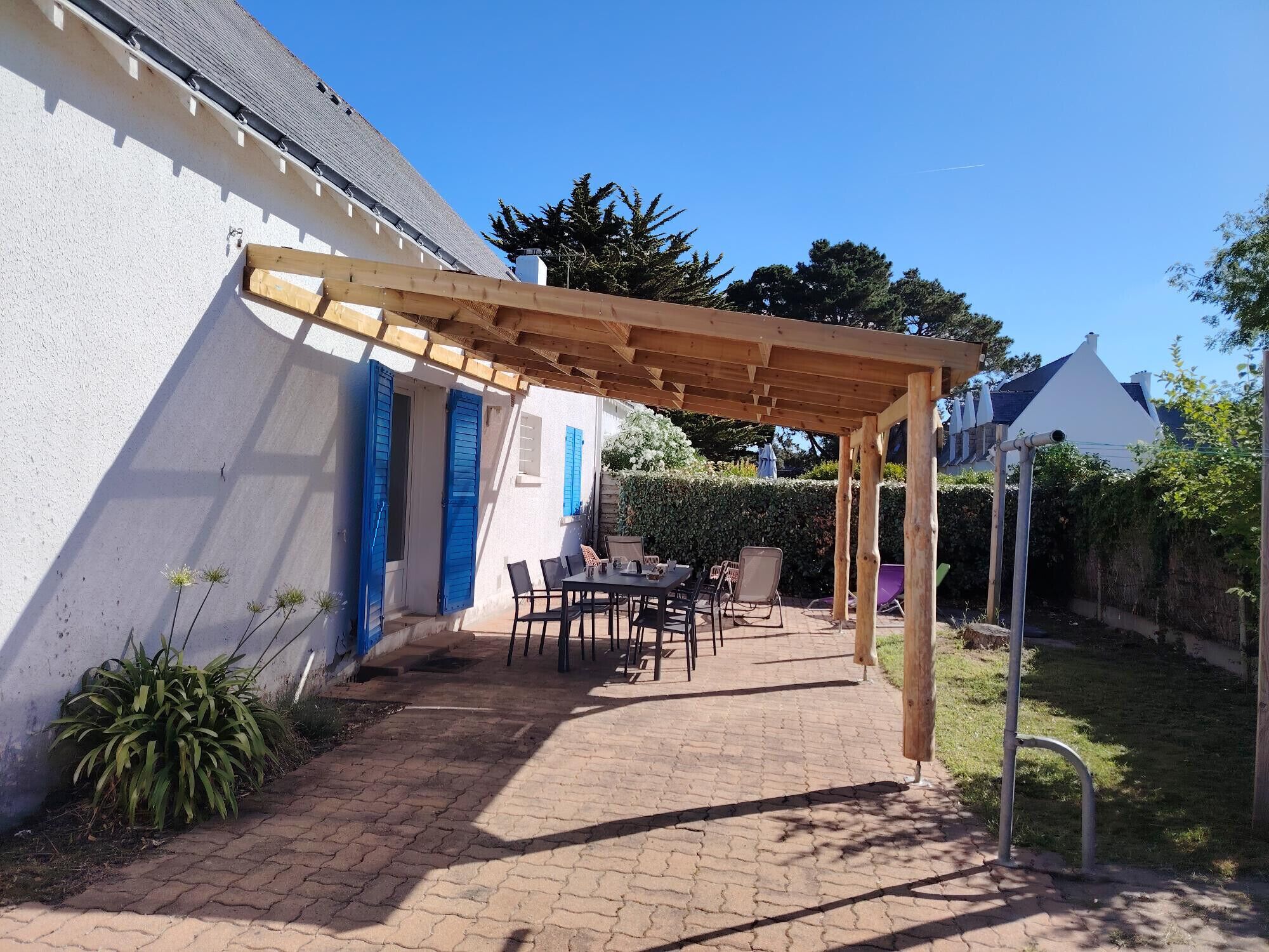 A covered patio area with a table and chairs, a wooden pergola, and a white building with blue shutters.