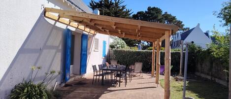 A covered patio area with a table and chairs, a wooden pergola, and a white building with blue shutters.