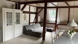 A loft bedroom with exposed wooden beams, a grey sofa, a white table, and a window with a view of greenery.