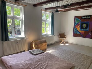 A room with a bed, a wicker chest, a folding chair, and a large window. - Stylish and bright apartment in a listed half-timbered house with a large garden (Lüchow)