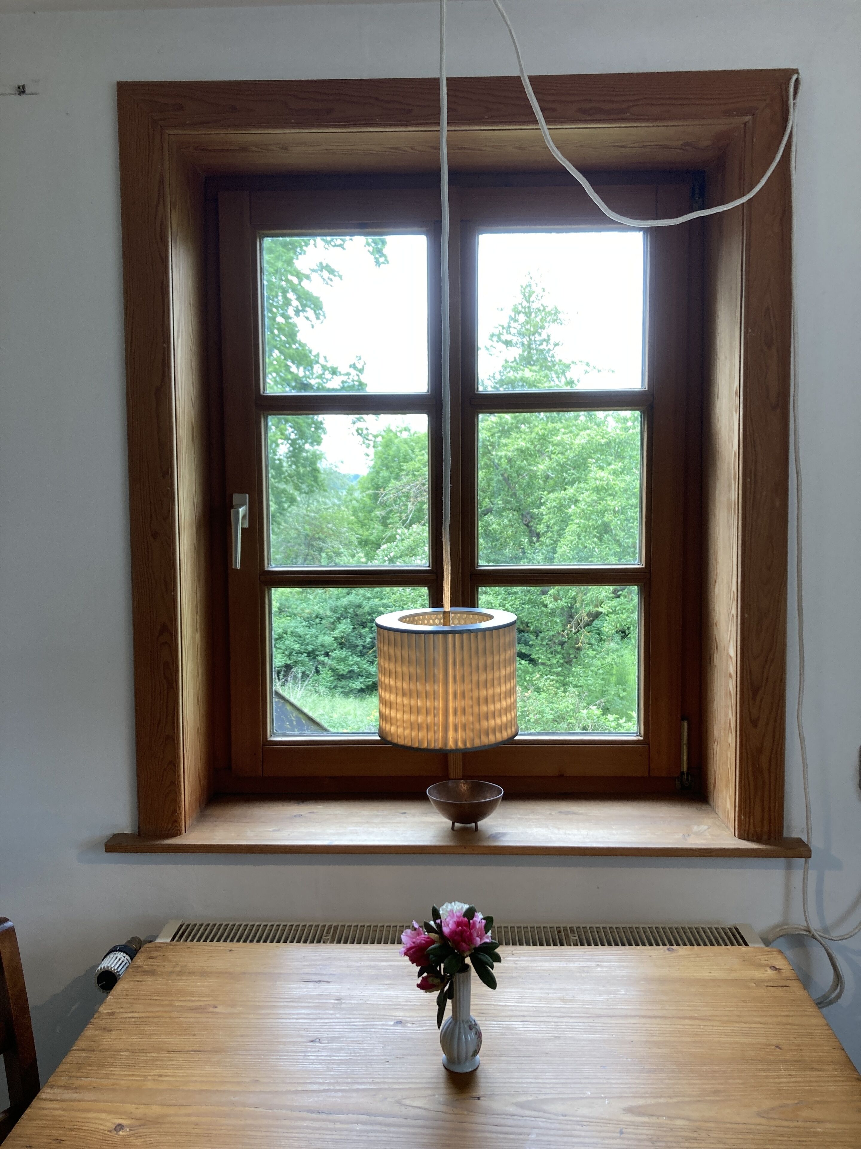 Et trævindue med en hængende lampe og en vase med blomster på et bord nedenunder.