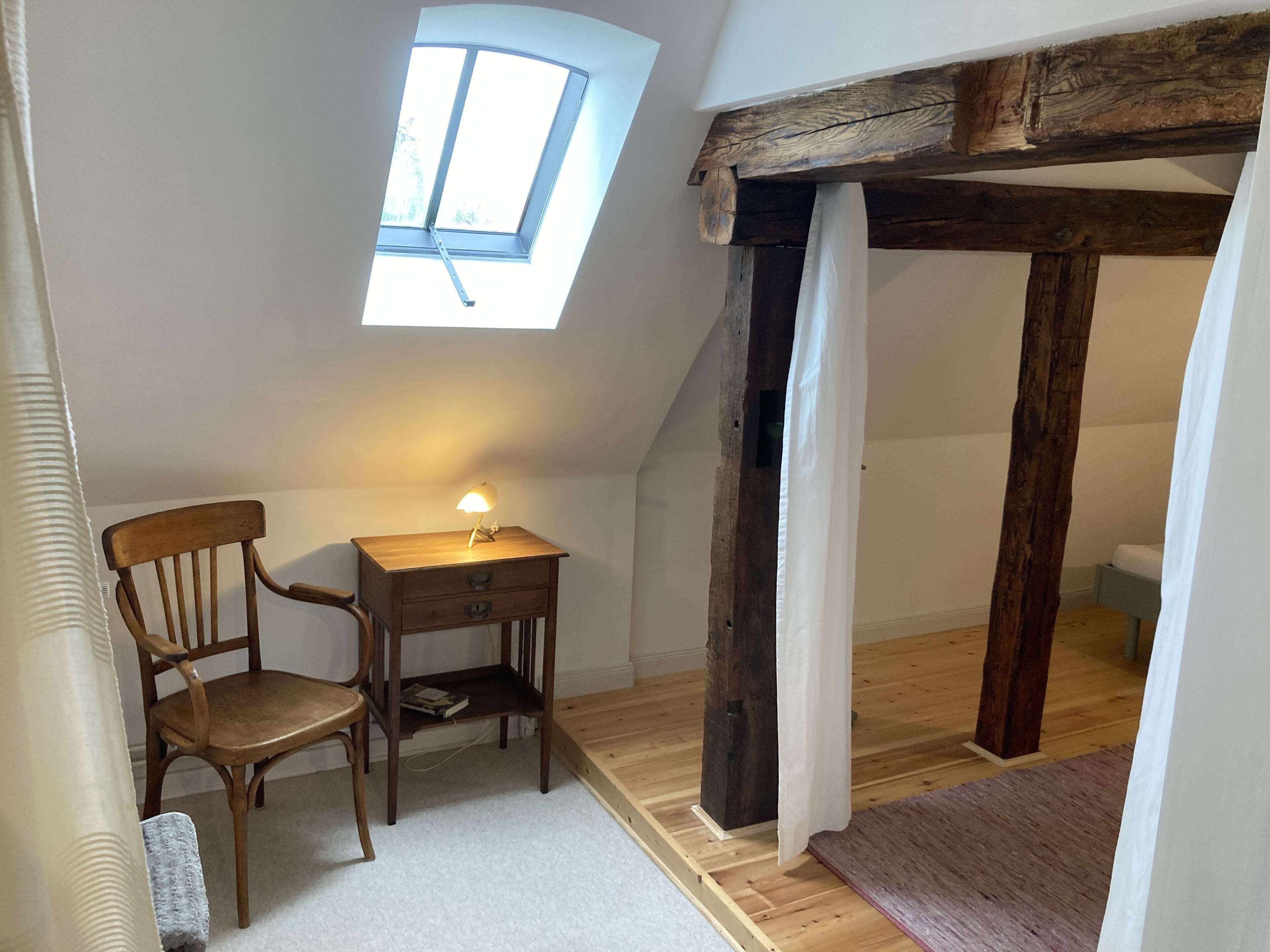 Een gezellig slaapkamer op zolder met dakraam, houten balken, een bureau met lamp en een stoel.