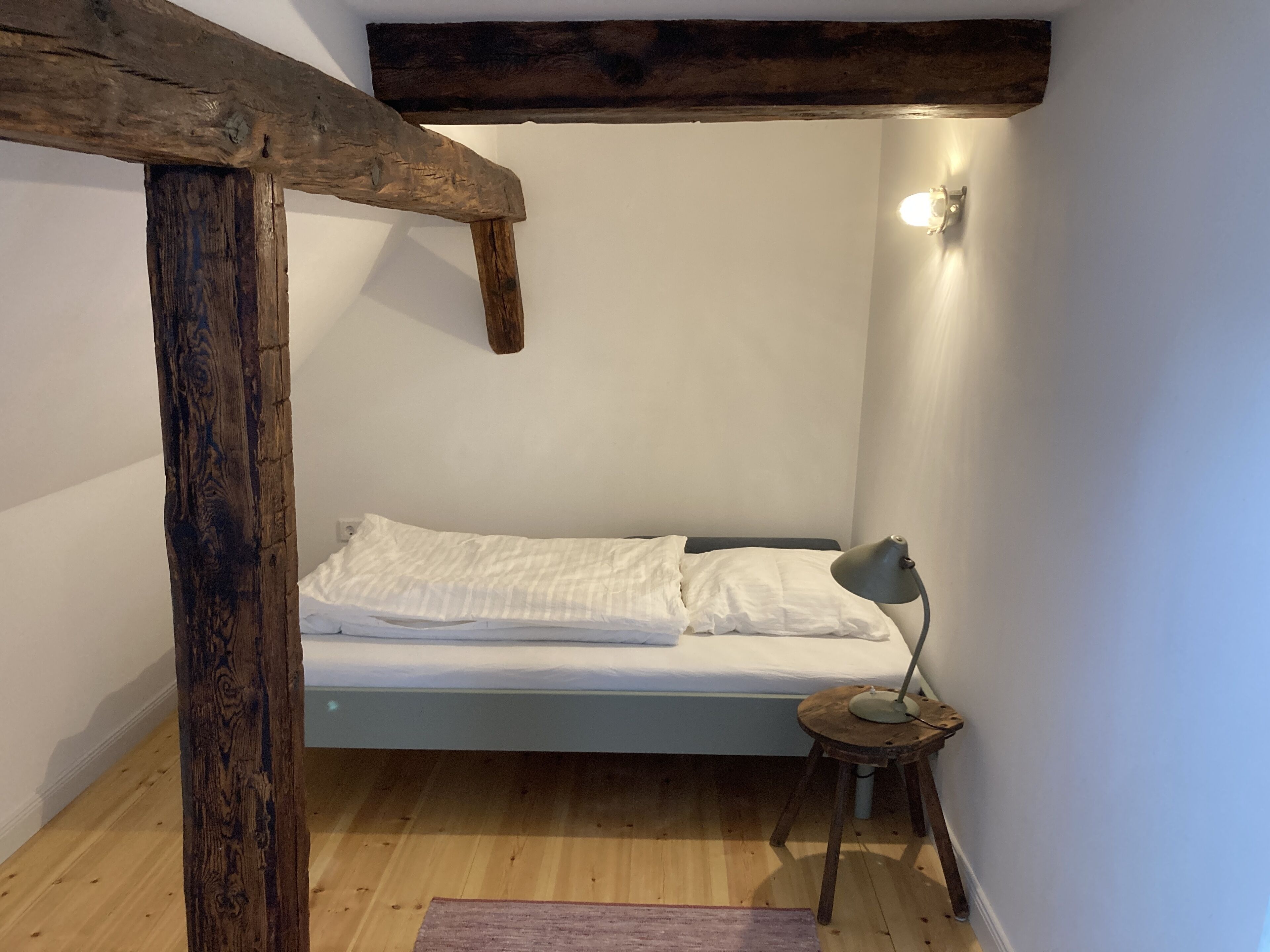 Een kleine kamer met een enkel bed, een houten balkenplafond, een bedlampje en een lichtschakelaar aan de muur.