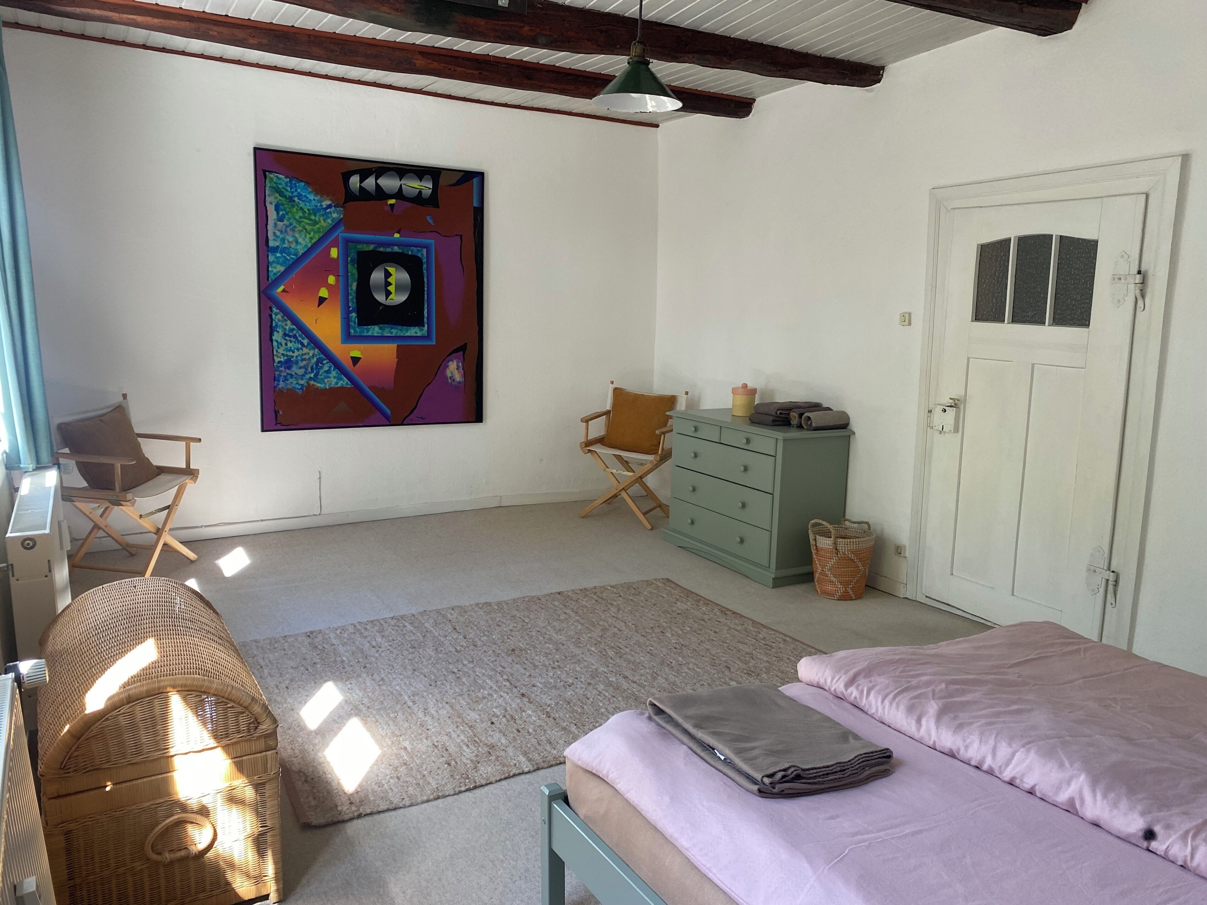 Une chambre à coucher avec un lit, deux chaises, une commode, une porte et un tableau abstrait aux couleurs vives accroché au mur.