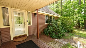 A house with a covered porch, a front door with a glass panel, and a small garden area with bushes and a wooden deck.