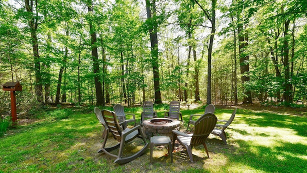 A circular fire pit surrounded by six Adirondack chairs in a forest setting.