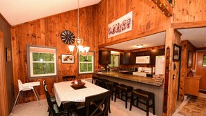 A wooden interior with a dining table, chairs, a kitchen area, and a view of trees through the windows.