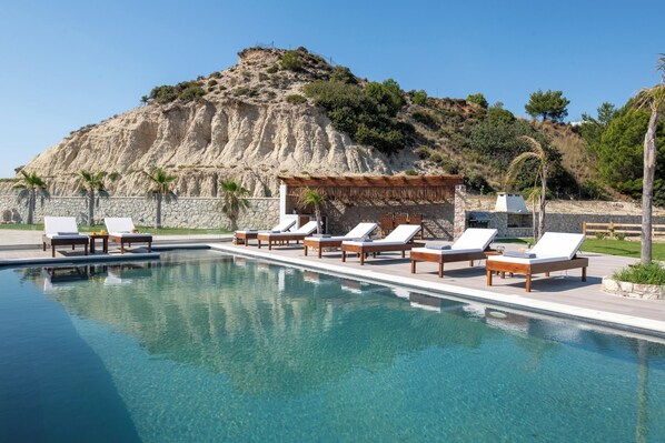 A pool with lounge chairs, a stone wall, and a mountain backdrop. - Waves of Blue Villa with Sea View (Tsairi)
