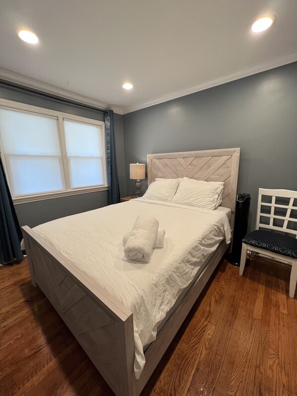 Una cama bien hecha con una toalla enrollada, una lámpara de mesita de noche y una silla en una habitación con suelo de madera y una ventana con cortinas.