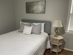 A neatly made bed with a grey headboard, a bedside table with a lamp, and a wall-mounted abstract art piece.