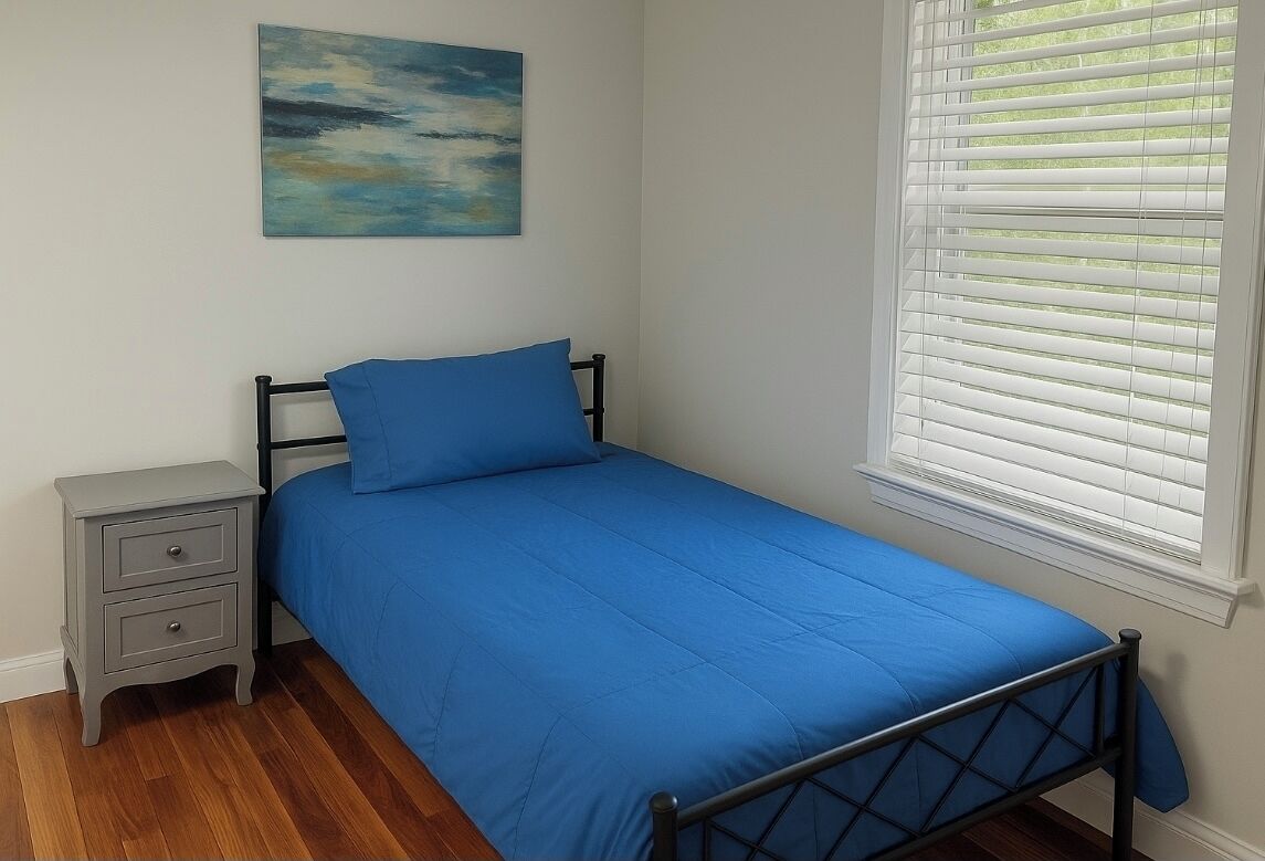 Een slaapkamer met een enkel bed, een nachtkastje met lades, een schilderij aan de muur en een raam met jaloezieën.