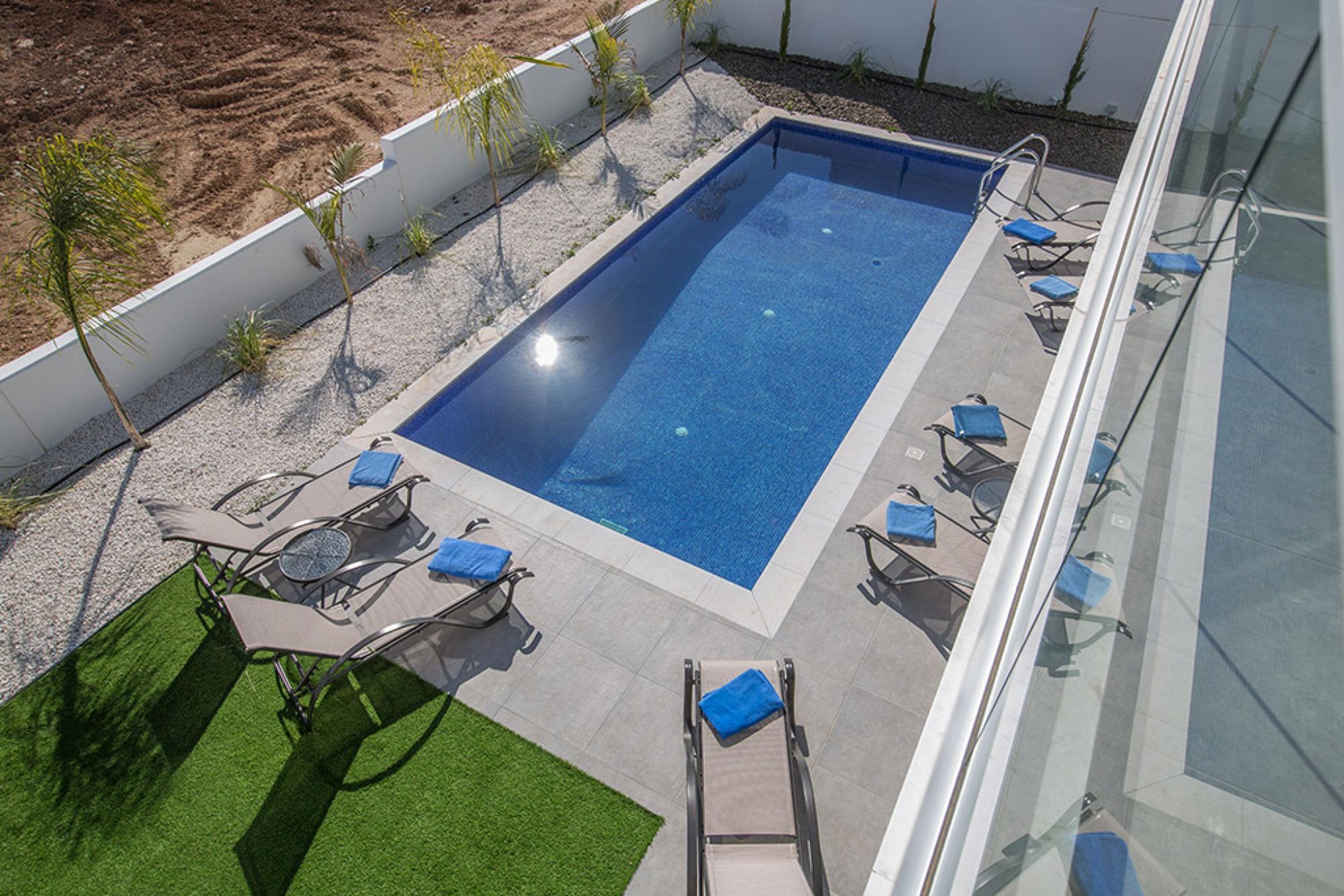 A rectangular outdoor swimming pool with a tiled deck, surrounded by lounge chairs and small palm trees.