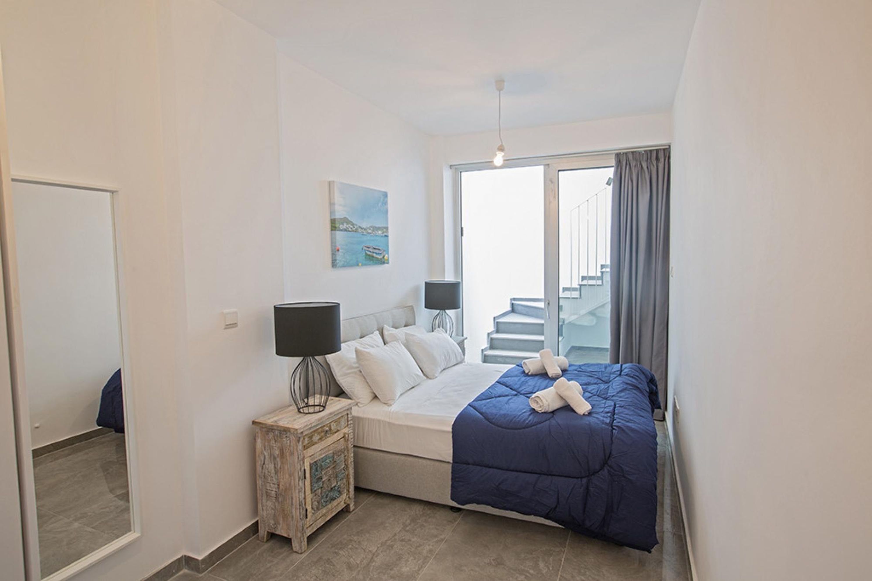 A bedroom with a bed, bedside tables, a painting, and a view of a staircase.