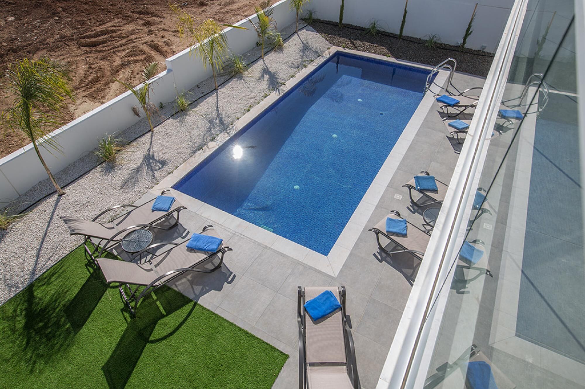 Une piscine rectangulaire à l’eau d’un bleu limpide, entourée d’un mur blanc et d’un espace recouvert de gravier où sont plantées de petites plantes. Plusieurs chaises longues avec des coussins bleus sont disposées autour de la piscine.