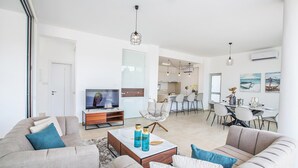 Un salon moderne avec un canapé, une table basse et un téléviseur. Il y a également un coin repas avec des chaises et une table. La pièce est dotée de murs blancs et d’une grande fenêtre.