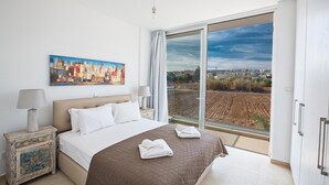 Une chambre d’hôtel avec un grand lit, un tableau représentant un paysage urbain, une vue sur un champ et une salle de bain moderne.