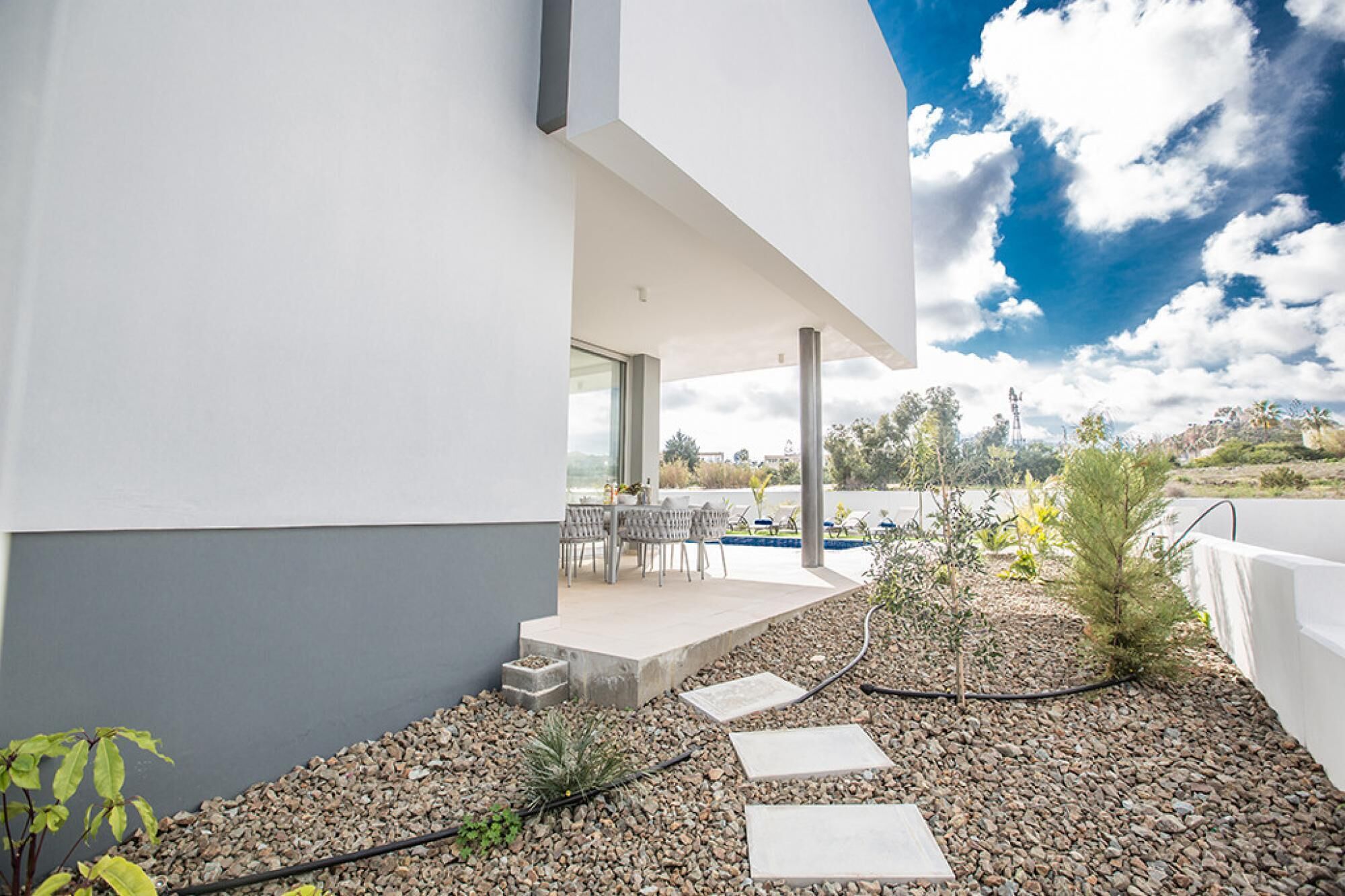 Un patio exterior moderno con una mesa y sillas, rodeado por un camino de grava y plantas pequeñas.
