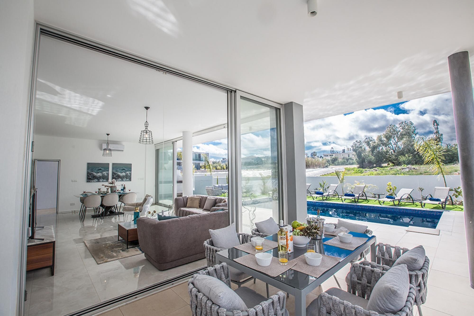 Una sala moderna con pared de vidrio, una piscina y mobiliario de exterior.