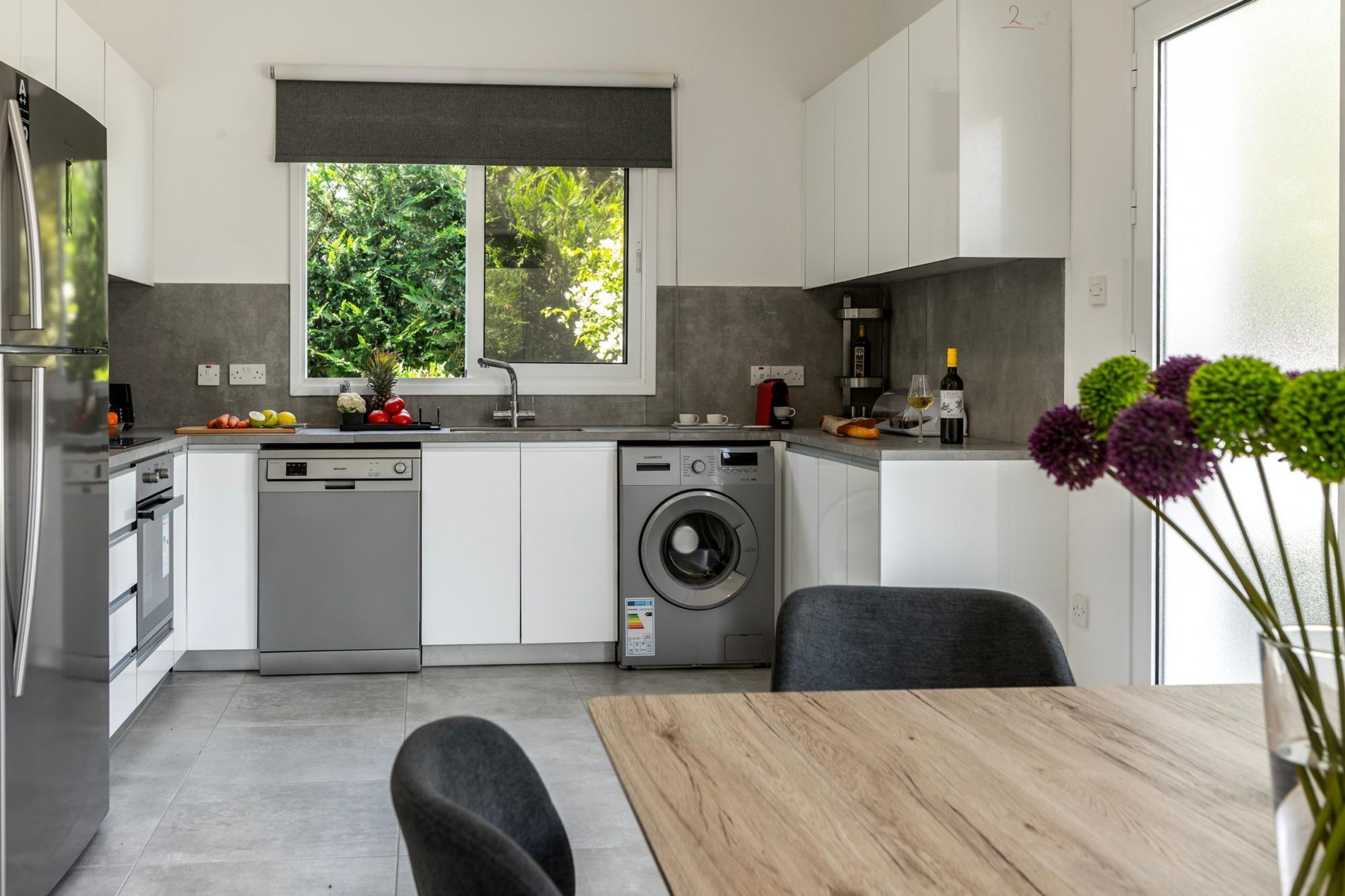 A modern kitchen with a dishwasher, washing machine, and a window with a view of greenery.