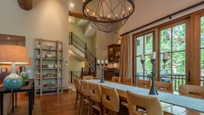 A spacious dining area with a long wooden table and chairs, a chandelier, and a staircase leading to an upper level.