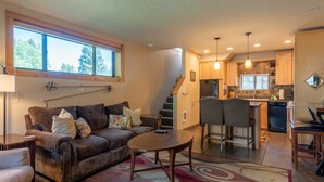 A cozy living room with a brown leather sofa, a coffee table, and a patterned rug. A staircase leads to a kitchen area with wooden cabinets and a dining table. - Alpine Villa Condo 1- Find a Perch in Ketchum at Treehouse Condo (Ketchum)