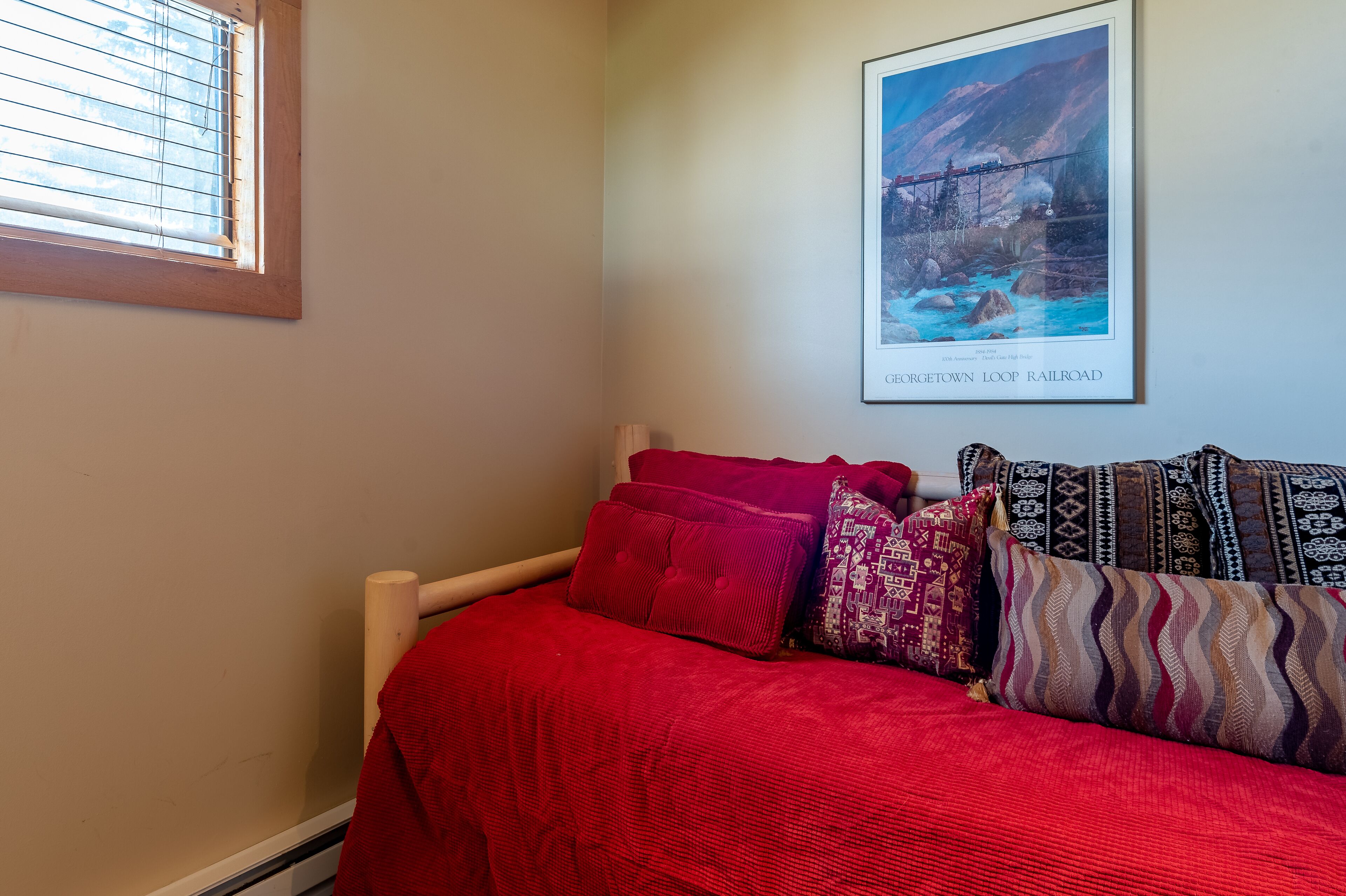 A bedroom with a wooden bed, red bedding, patterned pillows, a framed picture of a bridge, and a window with blinds.