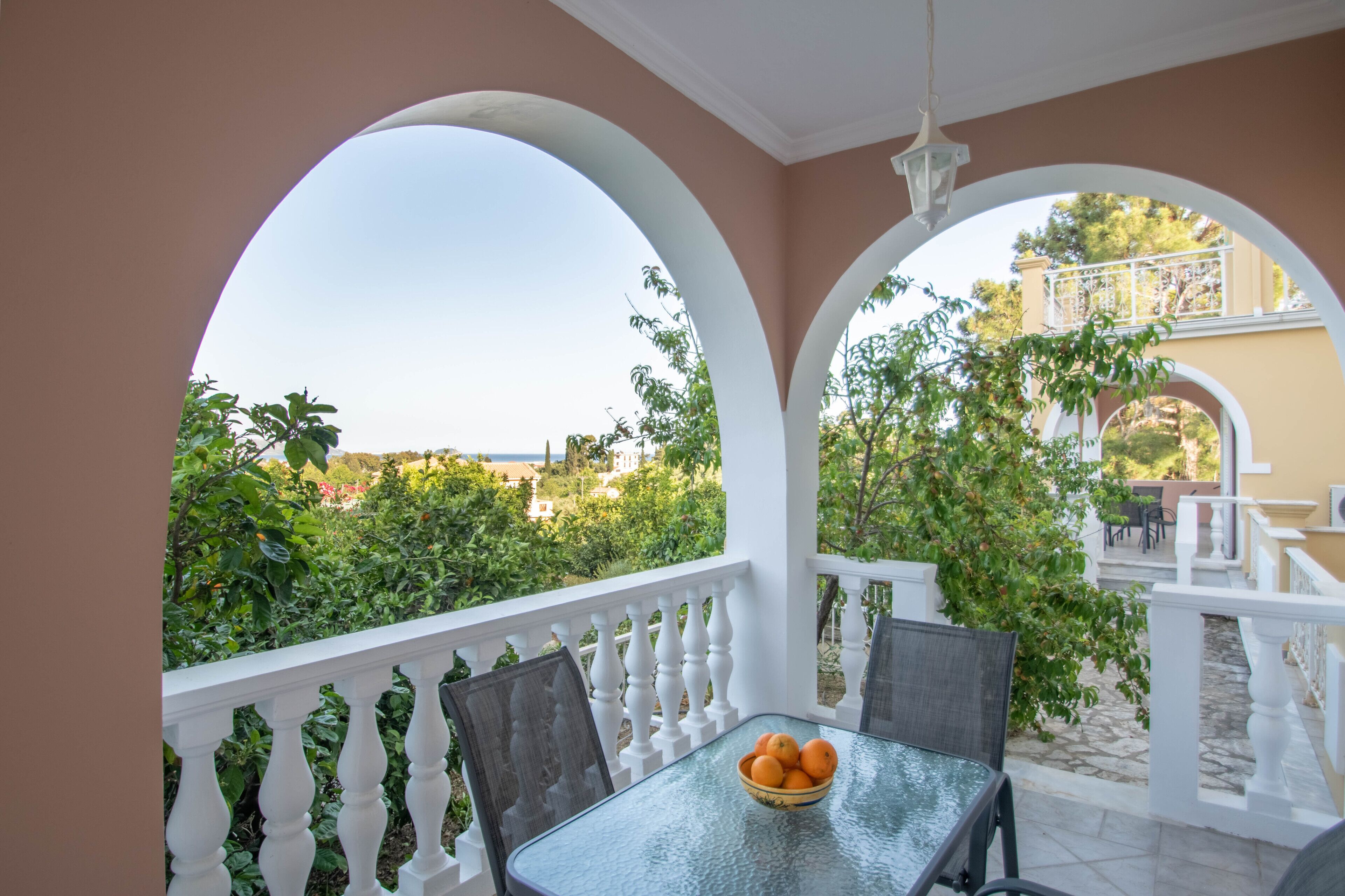 Varanda com mesa e cadeiras, uma tigela de frutas e vista para o verde e edifícios.