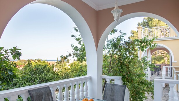 A covered outdoor seating area with a table and chairs, a bowl of oranges, and a view of greenery and buildings in the background. - Andelica Holiday Villa (Zakynthos)