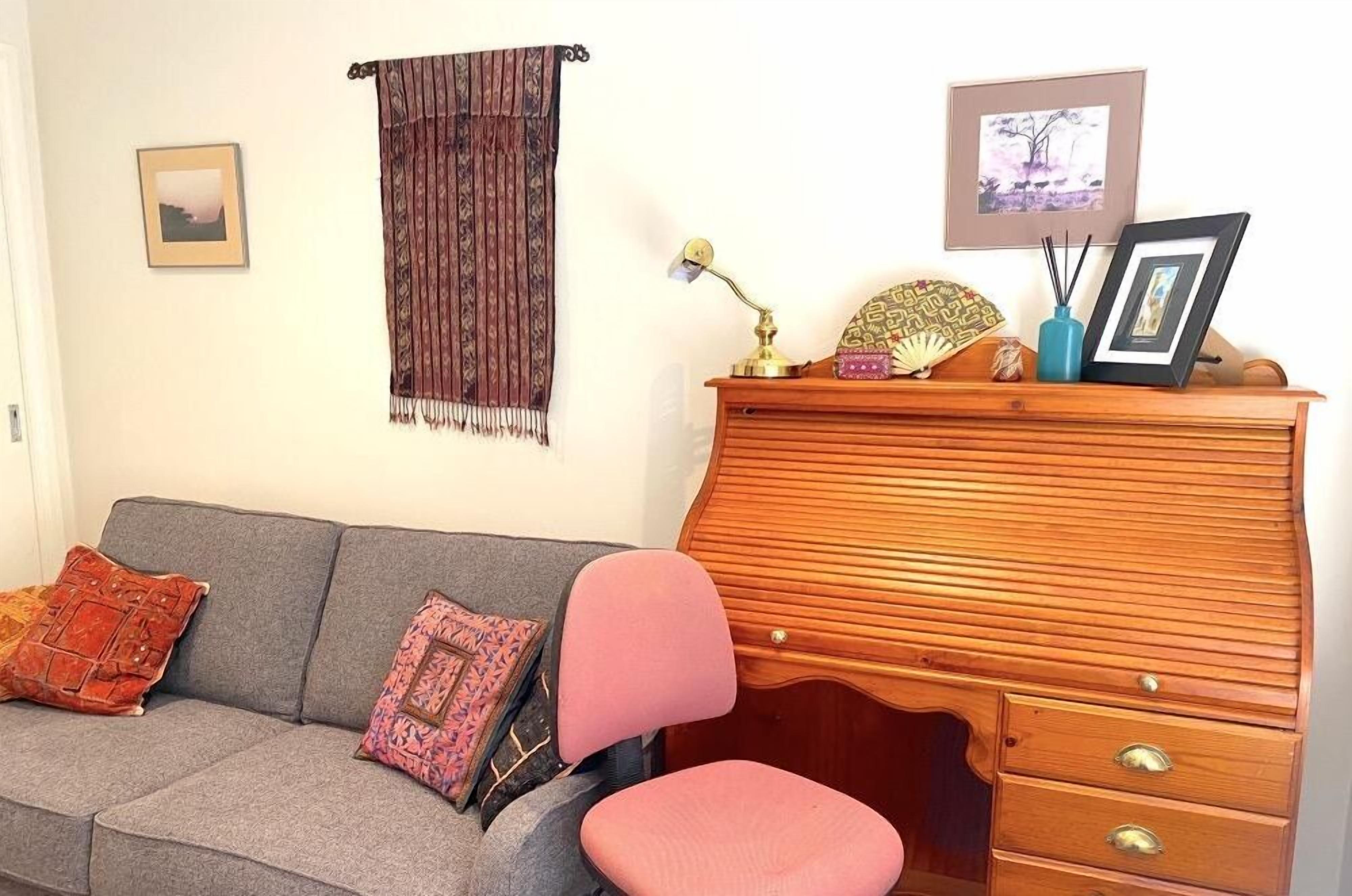 Een woonkamer met een grijze bank, een roze fauteuil, een houten kast met lades, een wandtapijt en ingelijste foto's aan de muur.