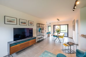 Ein modernes Wohnzimmer mit einem Flachbildfernseher, einer Holzkommode, einem Couchtisch mit Glasplatte und einem Essbereich mit einem gelben Tisch und blauen Stühlen.