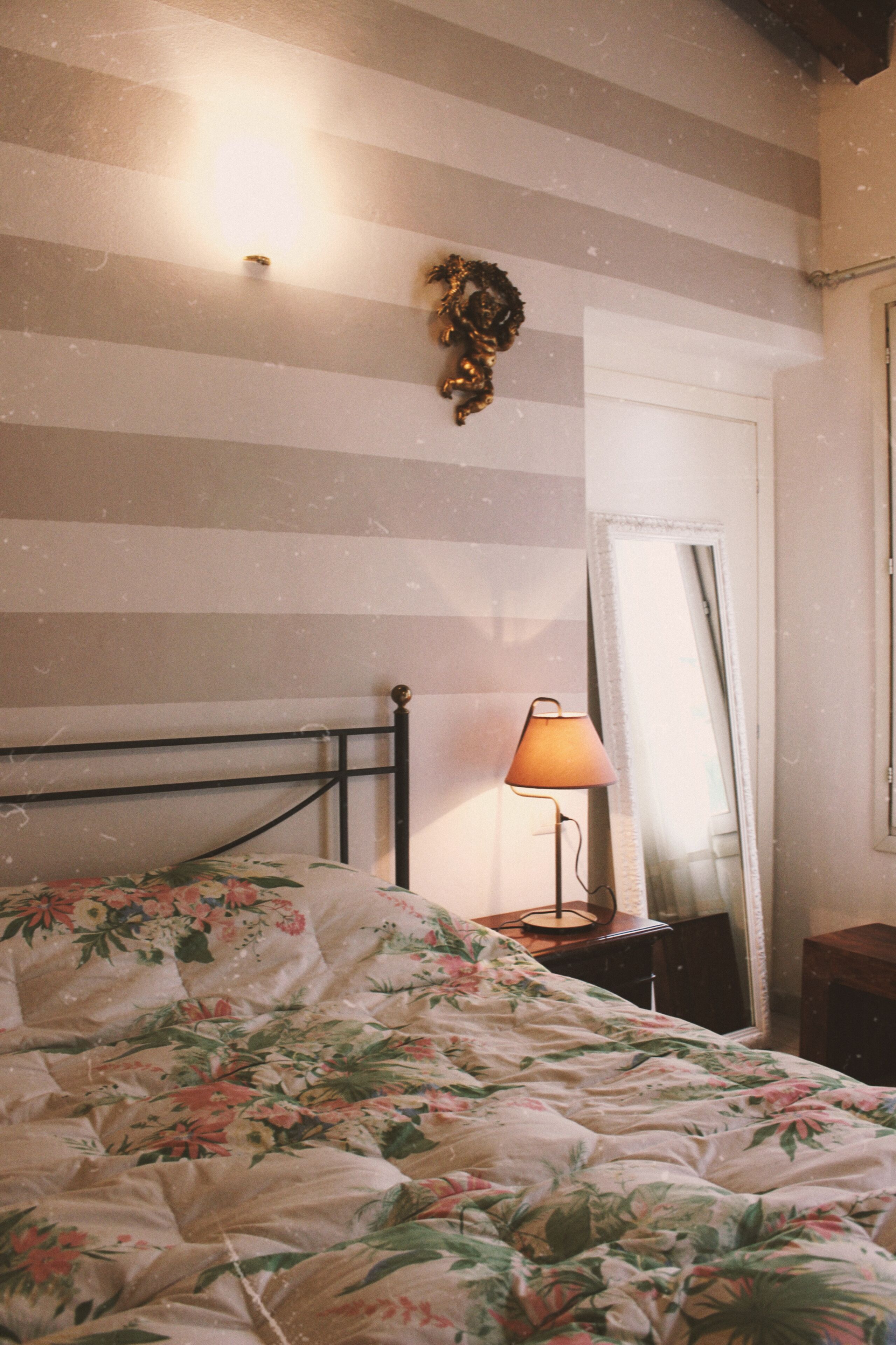 Una cama con ropa de cama floral, una lámpara de noche, un espejo y una pared con rayas horizontales.