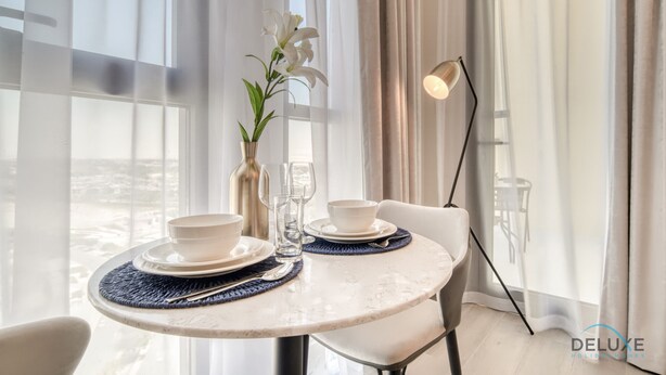 A dining area with a round table set for two, a vase with a plant, and a floor lamp. - Prime Studio at Mesk 1 Midtown Dubai Production City by Deluxe Holiday Homes (Dubai)