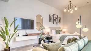 A modern hotel room with a flat-screen TV, a large mirror, a bed with white bedding, a green sofa, and a small side table with a lamp.