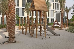 Un terrain de jeux avec un toboggan en bois et des structures d’escalade, entouré de palmiers et avec un bâtiment en arrière-plan.