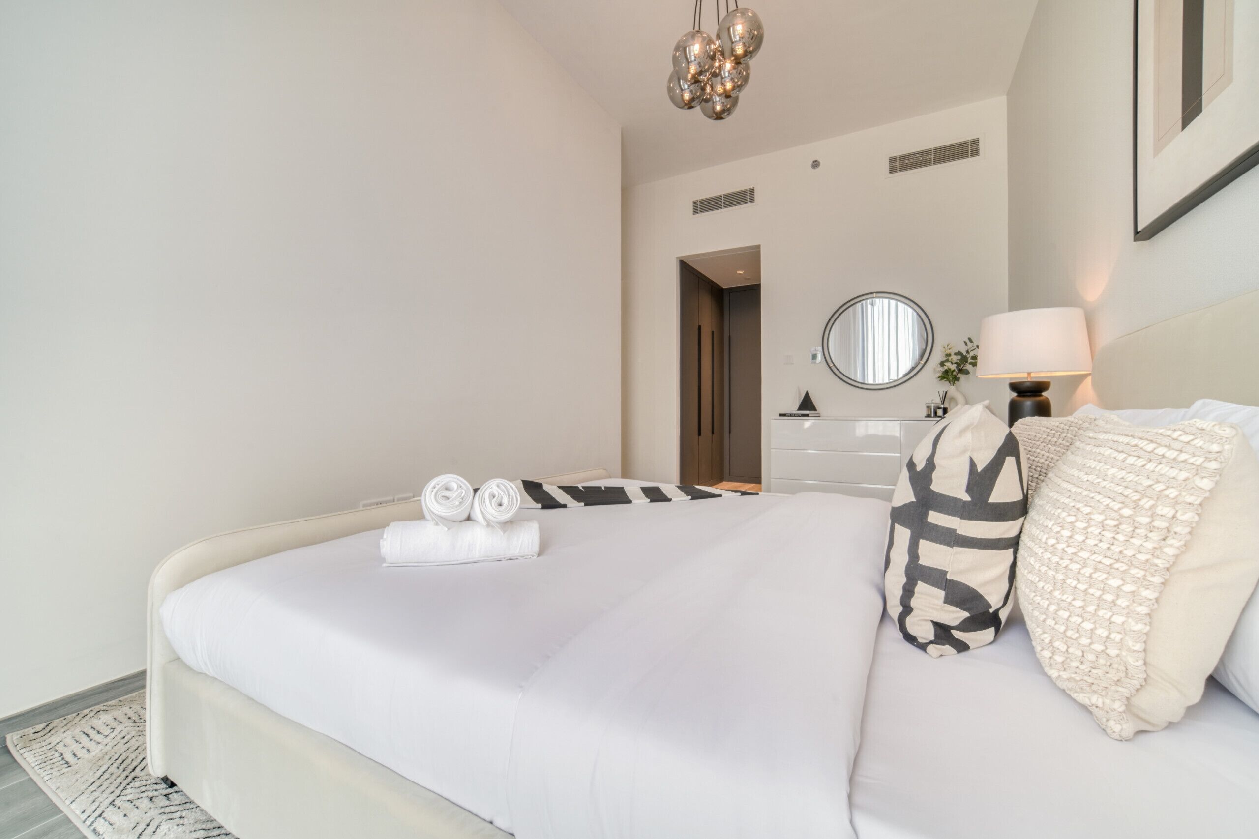 Una cama bien hecha con ropa blanca y dos toallas enrolladas, una lámpara de noche y un espejo en la pared.