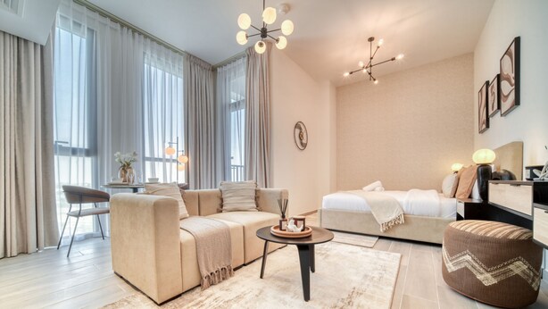 Una sala de estar moderna y bien iluminada, con un sofá, una mesa de centro, un reposapiés y una cama con cabecera.