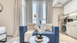 Ein modernes Wohnzimmer mit einem blauen Sofa, einem runden Couchtisch und Blick auf eine Küche mit einer Waschmaschine.