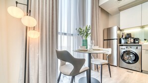A modern kitchen with a washing machine, a dining area with a table and chairs, and a large window with curtains. - Classic Studio at Mesk 1 Midtown Dubai Production City by Deluxe Holiday Homes (Dubai)